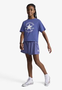 Μπλε κοντομάνικο t-shirt με λευκό αστέρι και κείμενο, συνδυασμένο με ταιριαστά μπλε σορτς και λευκά ψηλά αθλητικά παπούτσια με πολύχρωμες λεπτομέρειες.