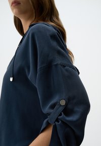 Maglietta leggera blu navy con vestibilità rilassata, dettaglio ai polsini con un bottone argentato e scollatura con cordoncino, realizzata in tessuto morbido.