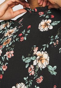 Schwarze Blumenbluse mit strukturiertem Finish. Enthält große cremefarbene und rosa Blumen mit grünen Blättern sowie ein Rüschenkragen-Detail.