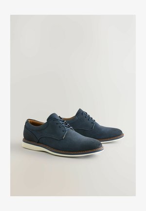Granatowe zamszowe buty sznurowane z zaokrąglonym noskiem, białą gumową podeszwą i kontrastującymi brązowymi przeszyciami na górnej krawędzi.