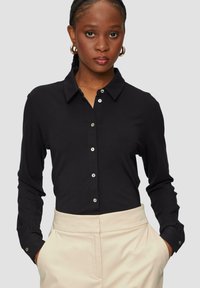 Black long-sleeve button-up shirt with a collar, featuring silver buttons and green button accents at the cuffs, paired with light beige trousers.