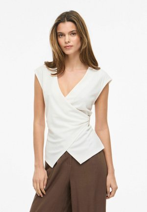 Młoda kobieta z prostymi brązowymi włosami, ubrana w białą bezrękawnikową bluzkę z wiązaniem i szerokie brązowe spodnie z wysokim stanem, stojąca na jednolitym tle.