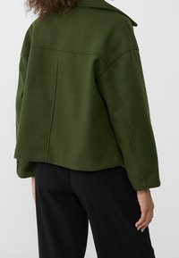 Mörkgrön oversize-jacka med krage, synlig sömnad och kortad fåll. Tillverkad av ett mjukt material, bärs över svarta byxor.