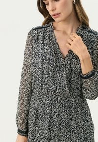 Bluza navy cu imprimeu floral, cu mâneci lungi translucide, guler cu volane și talie strânsă. Prezintă modele florale albe intricate și accente din dantelă.