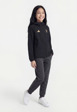 Fille souriante debout, portant une veste zippée noire Juventus, un jogging gris foncé et des baskets blanches sur fond blanc uni.