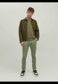 Olivově zelená bunda přes světle zelenou košili, v kombinaci s úzkými světle zelenými kalhotami a černobílými teniskami. Jednoduchý design, ležérní vzhled.