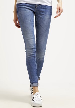 Personne portant un jean skinny bleu retroussé et des baskets blanches avec des bandes noires, marchant vers l'avant sur un fond uni.