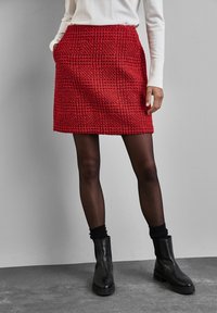 Röd texturerad kort kjol med ett rutmönster, utrustad med fickor, parat med svarta ankelskogs och genomskinliga svarta strumpbyxor.