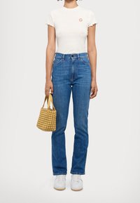 Bílý, krátký rukáv, texturovaný top s malým červeným logem; modré džíny s vysokým pasem; žlutá tkaná kabelka; bílé tenisky.