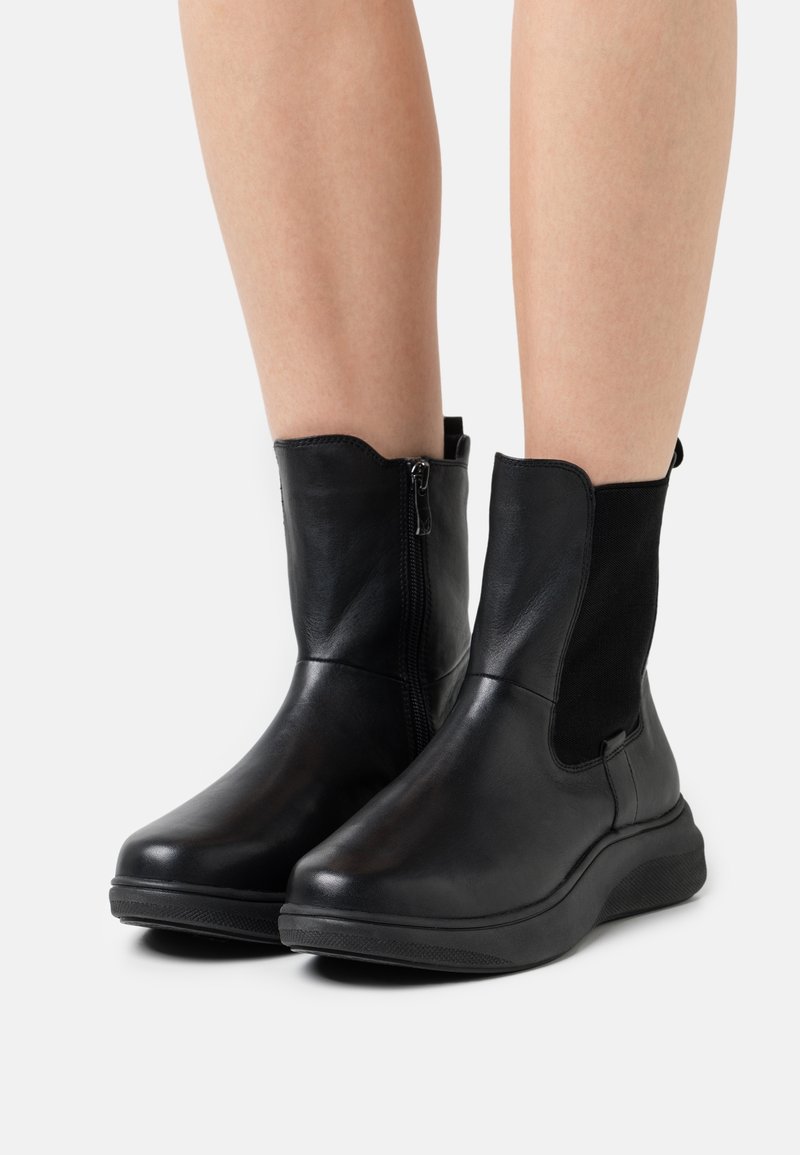 Bottes en cuir noir mi-mollet avec fermeture éclair latérale sur la botte gauche et panneau élastique sur la botte droite, portées sur des jambes nues sur fond blanc.