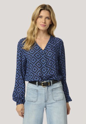 Blouse bleue à motifs avec un design géométrique et des accents sombres, décolleté en v, et manches longues, assortie à un jean clair taille haute.