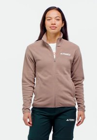 Braune Fleecejacke mit hohem Kragen und Reißverschluss; verfügt über das "TERREX"-Logo auf der linken Brust. Kombiniert mit dunkelgrünen Hosen.