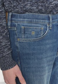 Personne portant un jean en denim bleu et un pull en maille gris foncé, main reposant près de la poche avant du jean.