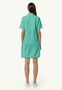 Grün gestreiftes Kleid mit kurzen Ärmeln, einem Kragen und einem gerafften Rock. Der Stoff wirkt leicht und glatt. Die Schuhe sind weiß mit silbernen Akzenten.