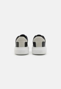Baskets en suède noir et gris avec une semelle en caoutchouc blanc, présentant une finition lisse et un branding minimal au niveau du talon. Vue arrière.