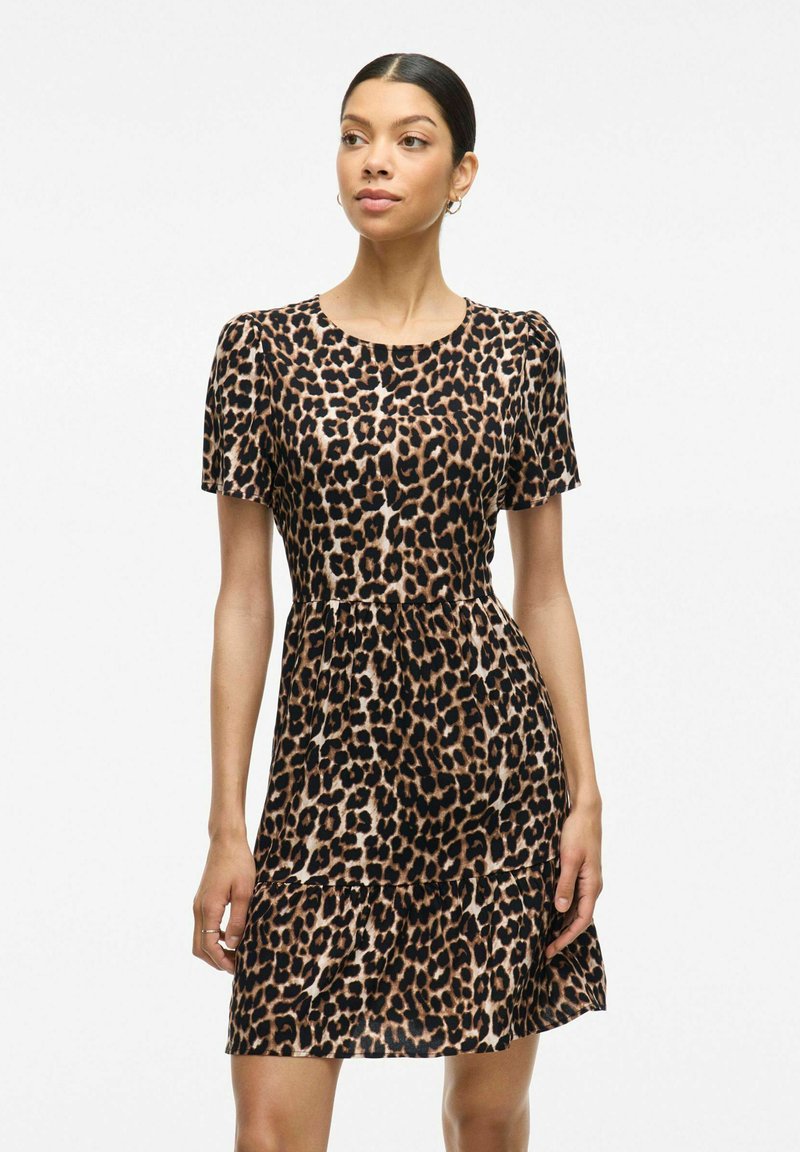 Šaty s leopardím vzorom, krátkymi rukávmi, okrúhlym výstrihom, viacerými volánmi na spodnej časti a ľahkým materiálom. Dominantné farby sú hnedá, čierna a béžová.