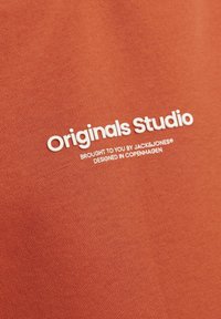 Narančasta tkanina s glatkom teksturom. Sadrži bijeli otisak teksta: "Originals Studio, donosi vam JACK&JONES, dizajnirano u Kopenhagenu."