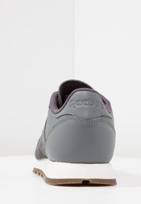 Chaussure de sport grise avec une tige en cuir et en daim, un col rembourré et une semelle en caoutchouc. Présente "Reebok" en relief sur le talon.