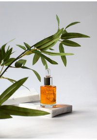 En genomskinlig glasflaska med ett silverdropparhuvud som innehåller en orange serum, placerad på en marmorplatta, omgiven av gröna blad.