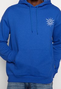 Sweat à capuche en coton bleu avec poche kangourou, capuche à cordon et motif soleil sur le devant. Comprend des poignets et un ourlet côtelés pour un ajustement confortable.