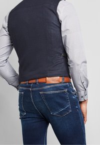 Mörkblå herringbone-väst, vit prydnadsskjorta, brun läderbälte och mörkblå jeans. Synlig sömnad och fickdetaljer på jeansen.