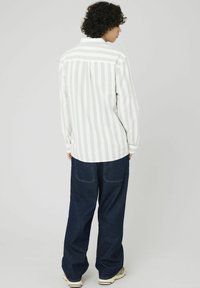 Personne avec des cheveux bouclés portant une chemise rayée blanche et beige, un jean large noir et des baskets blanches, se tenant de dos.