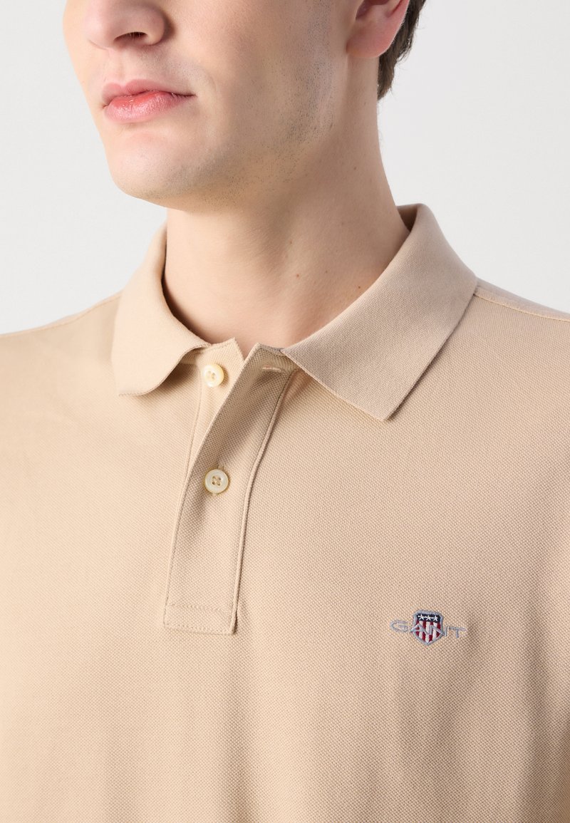 Beige Poloshirt aus strukturiertem Baumwollstoff, mit einer Knopfleiste mit zwei Knöpfen und einem gestickten Logo auf der linken Brust.