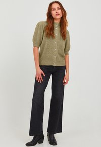 Olivno zeleno pleteno kratkoročno majico z visokim ovratnikom in zaponko na gumb, v kombinaciji s širokimi temnimi kavbojkami in črnimi gležnarji.