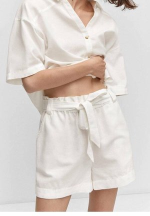 Femme portant un short taille paperbag blanc avec une ceinture en tissu nouée en nœud, associé à une chemise blanche à manches courtes et boutonnée.