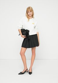 Sweat-shirt blanc avec motif de soleil, mini-jupe plissée noire et ballerines noires à bout pointu. Sac noir avec détail noué à la main.