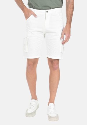 Uomo che indossa pantaloncini cargo bianchi con tasche laterali e sneakers bianche, in piedi con una mano in tasca davanti a uno sfondo semplice.