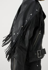 Zwarte leren jas met franjes langs de schouders, zilveren studs, een ritssluiting op de zak en een verstelbare riem. Gestructureerde afwerking.