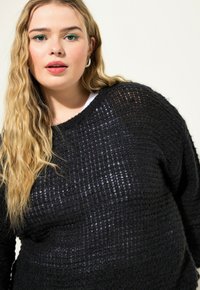 Schwarzer Strickpullover mit einem strukturierten, gewebten Design. Verfügt über einen weiten Halsausschnitt und einen lockeren Schnitt, kombiniert mit einer weißen inneren Schicht.