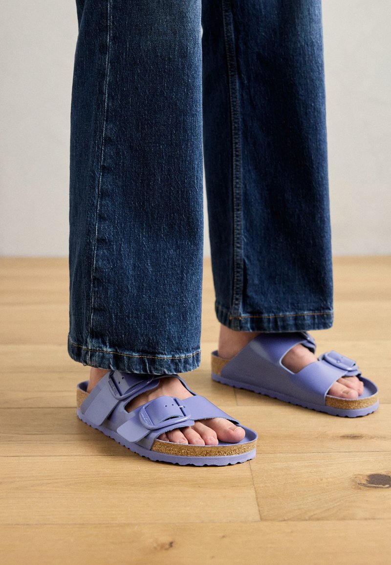 Lila Sandalen mit zwei verstellbaren Riemen, Korkfußbett und strukturiertem Gummisohle, getragen mit dunklen Jeans, auf einem Holzfußboden.