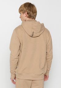 Beige hettegenser med snor i hetta, ribbestrikkede mansjetter og nederkant. Har en liten logo på baksiden av hetten. Mykt materiale og uformelt design.