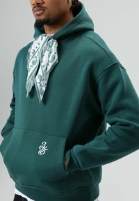 Groene trui met capuchon, met een witte paisley-bandana gestrikt bij de hals en een klein wit logo op het voorzakje.