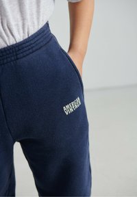 Tumši zili vilnas sporta bikses ar elastīgu jostu, sānu kabatām un balta izšūta logotipu uz augšstilba ar uzrakstu "AMERICAN VINTAGE".