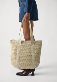 Grande borsa a tracolla in paglia intrecciata con texture beige e spalline piatte a contrasto. Su uno sfondo chiaro, tenuta in mano da una persona.