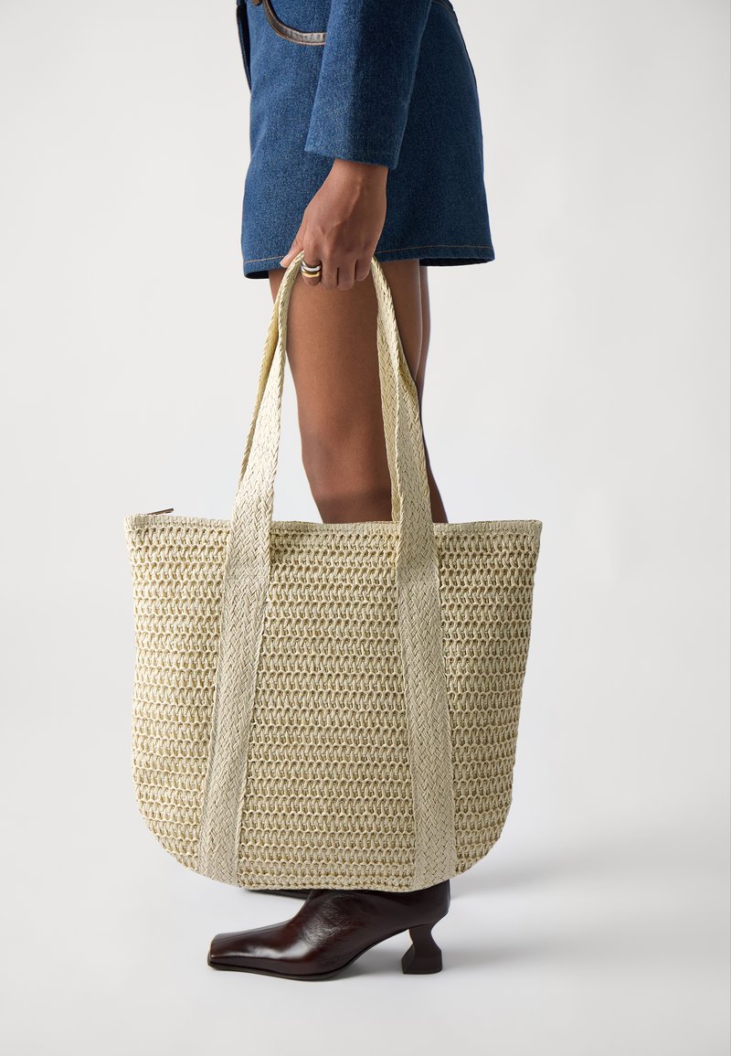 Grande borsa a tracolla in paglia intrecciata con texture beige e spalline piatte a contrasto. Su uno sfondo chiaro, tenuta in mano da una persona.