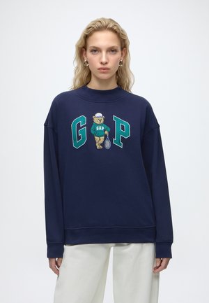 Ung kvinde med blondt hår bærer en mørkeblå sweatshirt med en tennisbjørn og store grønne bogstaver G og P, kombineret med hvide bukser.