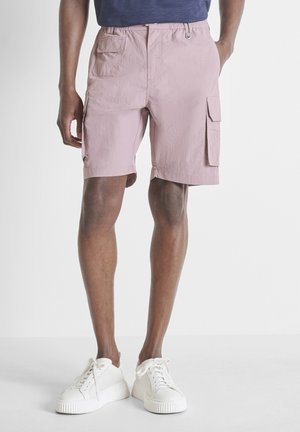 Hombre con pantalones cortos cargo de color rosa claro con múltiples bolsillos y zapatillas blancas sobre fondo claro liso.