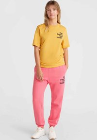 Gul bomullströja med svart text, i kombination med rosa sweatpants med en liknande tryck. Modellen bär vita sneakers.