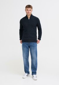 Marineblauer Strickpullover mit einem Halb-Reißverschluss-Kragen, kombiniert mit hellblauen Jeans und weißen Sneakers mit schwarzen Akzenten. Weiche Textur und lässige Passform.