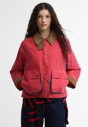 Młoda kobieta z długimi, kręconymi włosami, ubrana w czerwoną kurtkę z beżowym kołnierzem i dużymi przednimi kieszeniami, z rękami w kieszeniach, na jednolitym tle.