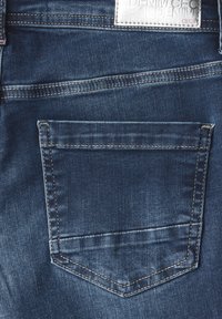Vaqueros de mezclilla azul oscuro con un bolsillo trasero único con costura curva y una etiqueta de tela justo encima del bolsillo. Textura suave.