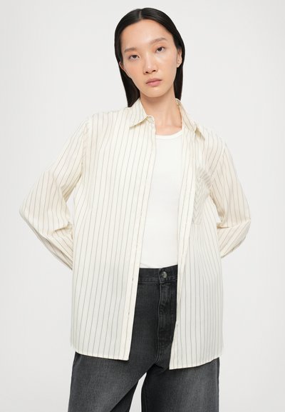Long-sleeved cream shirt with vertical black stripes, featuring a button-down collar and a pocket, paired with a white undershirt and dark jeans.