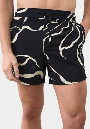 Sorte badeshorts med hvide abstrakte bølgemønstre, elastisk taljebånd og snor, båret af en person, der står med den ene hånd i lommen.