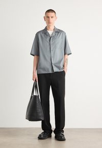 Camisa gris de manga corta con botones y bolsillo en el pecho, combinada con pantalones negros de corte recto y un bolso de cuero negro, llevada con zapatos negros.