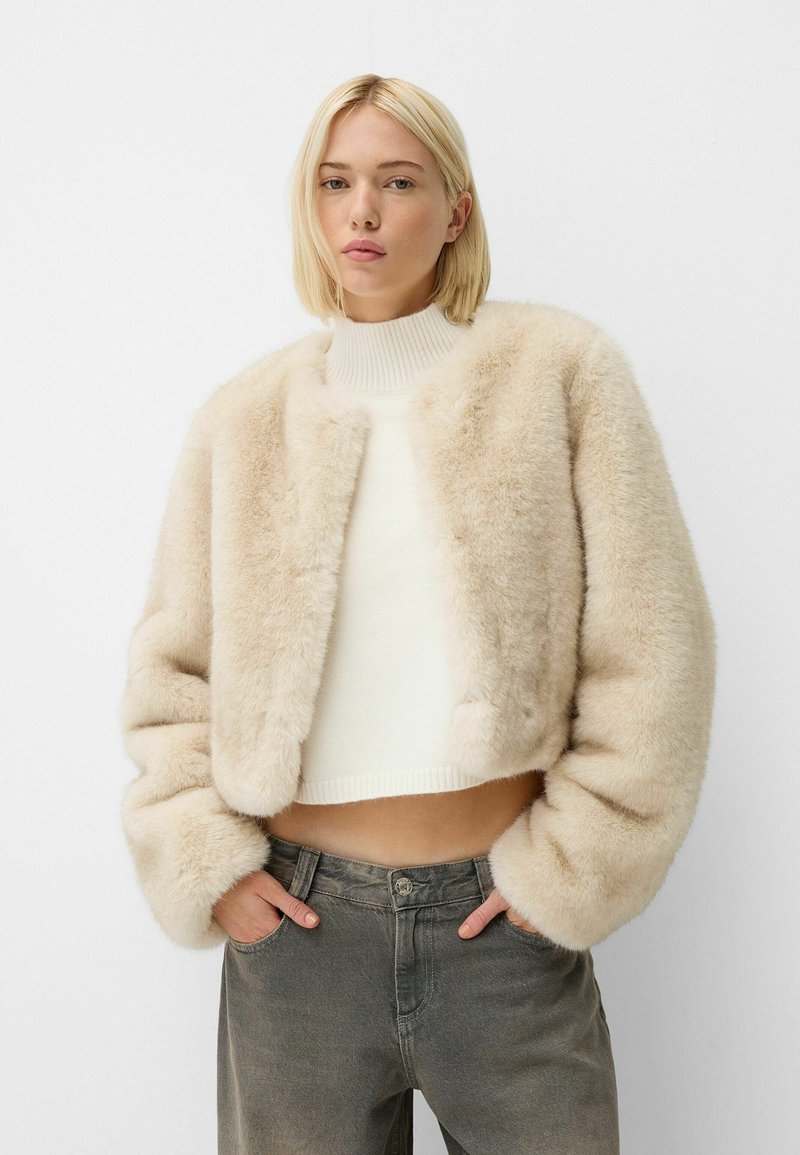 Giacca cropped in pelliccia sintetica beige, con colletto rotondo, texture morbida e maniche ampie, abbinata a un dolcevita a costine color crema.