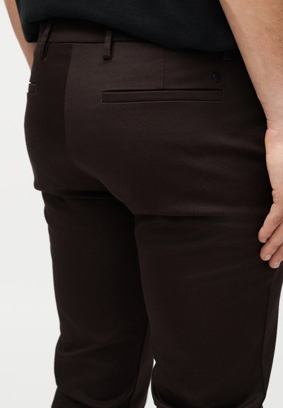 Gros plan sur le bas du dos d'une personne portant un pantalon ajusté marron foncé avec des poches passepoilées arrière et une chemise noire rentrée à l'intérieur.